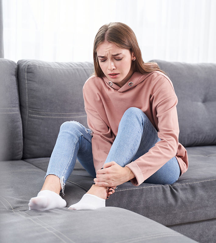 ankle pain woman sitting on a couch grabbing her ankle in pain